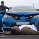 A man is lying in front of a damaged car which has just hit him. The responsible driver is standing at the door of the car, looking at him with a worried expression. A man is lying in front of a damaged car which has just hit him. The responsible driver is standing at the door of the car, looking at him with a worried expression.