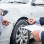 Insurance agent writing on clipboard while examining car after accident claim being assessed and processed. Insurance agent writing on clipboard while examining car after accident claim being assessed and processed.